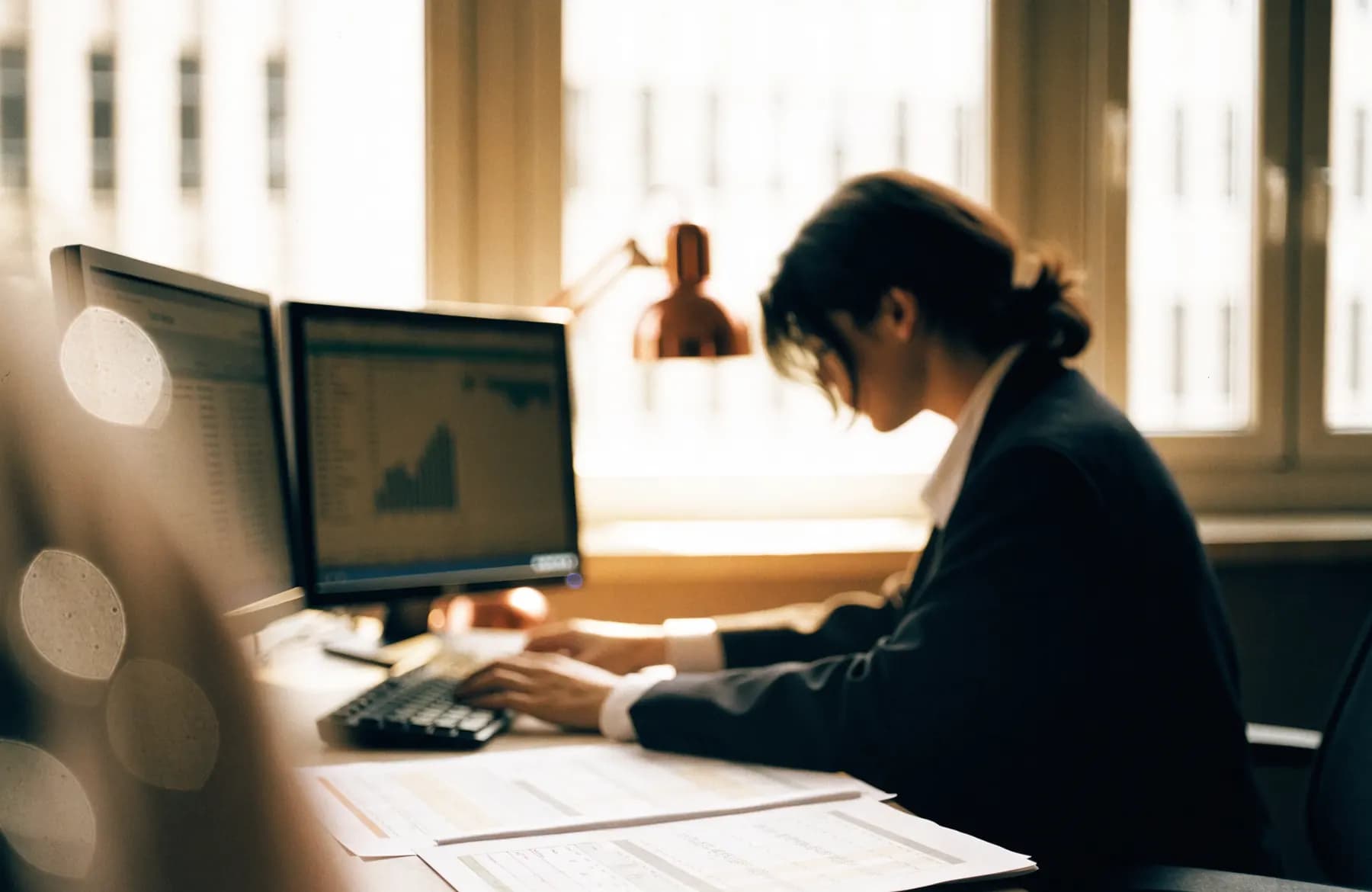 A financial analyst working across two monitors with printed reports beside the keyboard